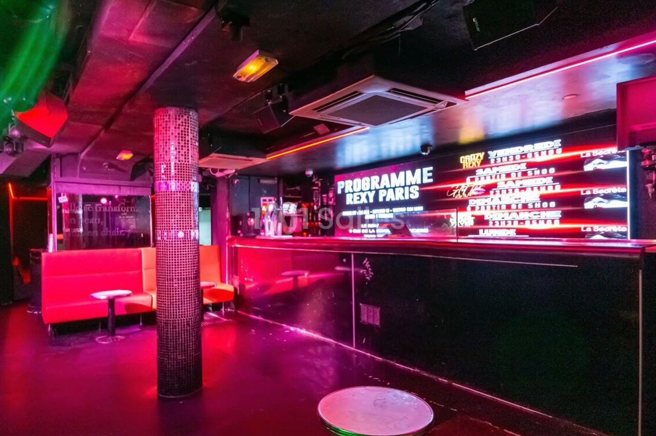 Intérieur d'une discothèque avec banquettes rouges, bar éclairé par des néons et colonne décorative au centre.