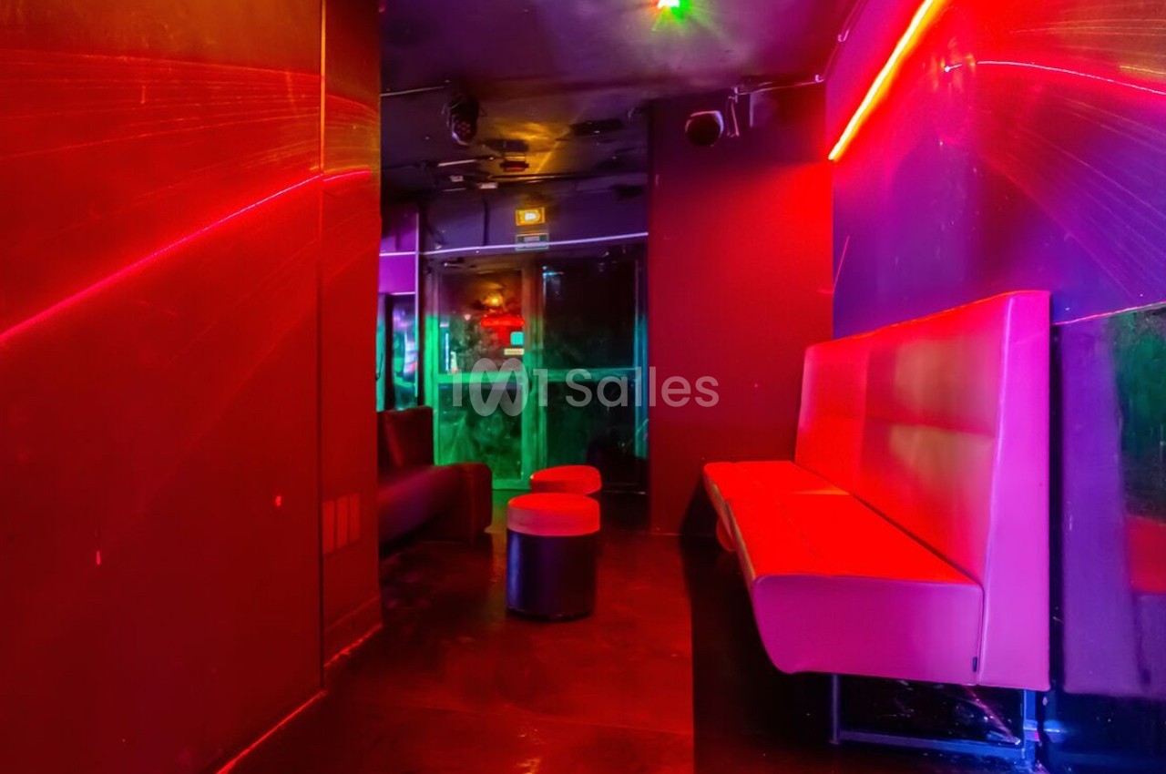 Salle de club avec éclairage néon rouge et violet, banquettes rouges et tabourets noirs sur un sol sombre.