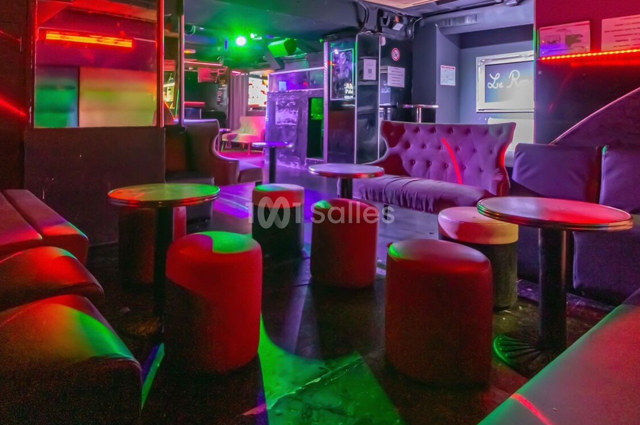 Salle de bar avec canapés, tabourets et tables basses, éclairée par des lumières colorées dans une ambiance tamisée.
