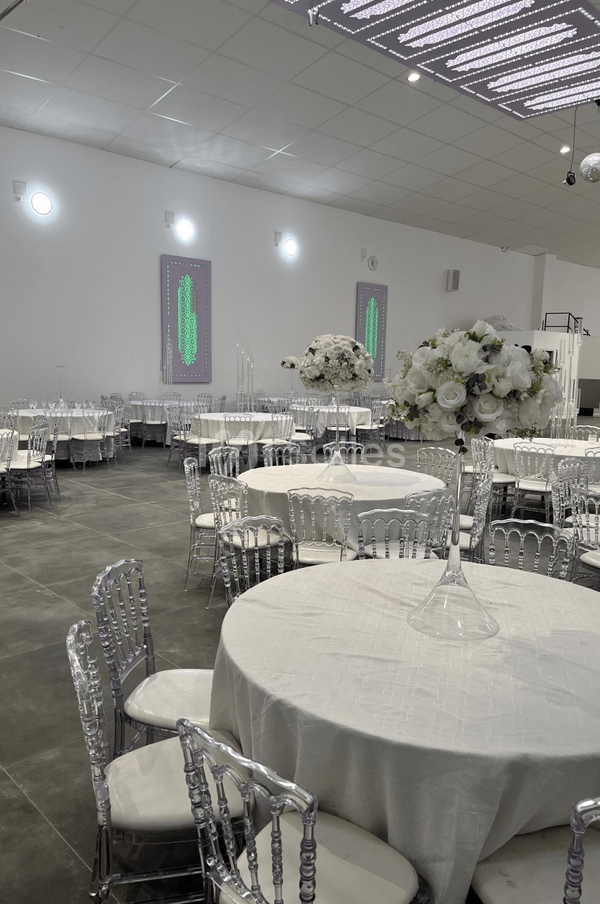 Salle de réception avec tables rondes dressées de nappes blanches, chaises transparentes et décorations florales.