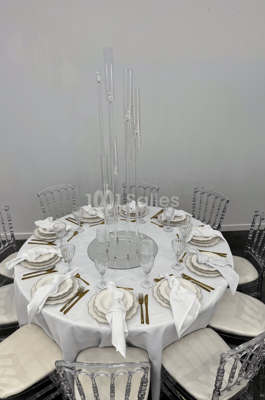 Table ronde dressée avec nappe blanche, couverts dorés, serviettes pliées et centre de table en tubes transparents.