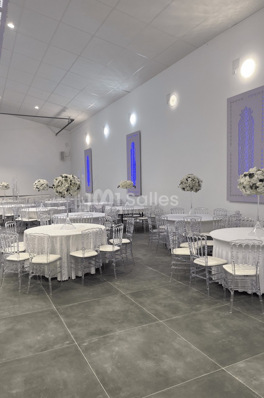 Salle de réception décorée avec des tables rondes, nappes blanches, chaises transparentes et arrangements floraux blancs.