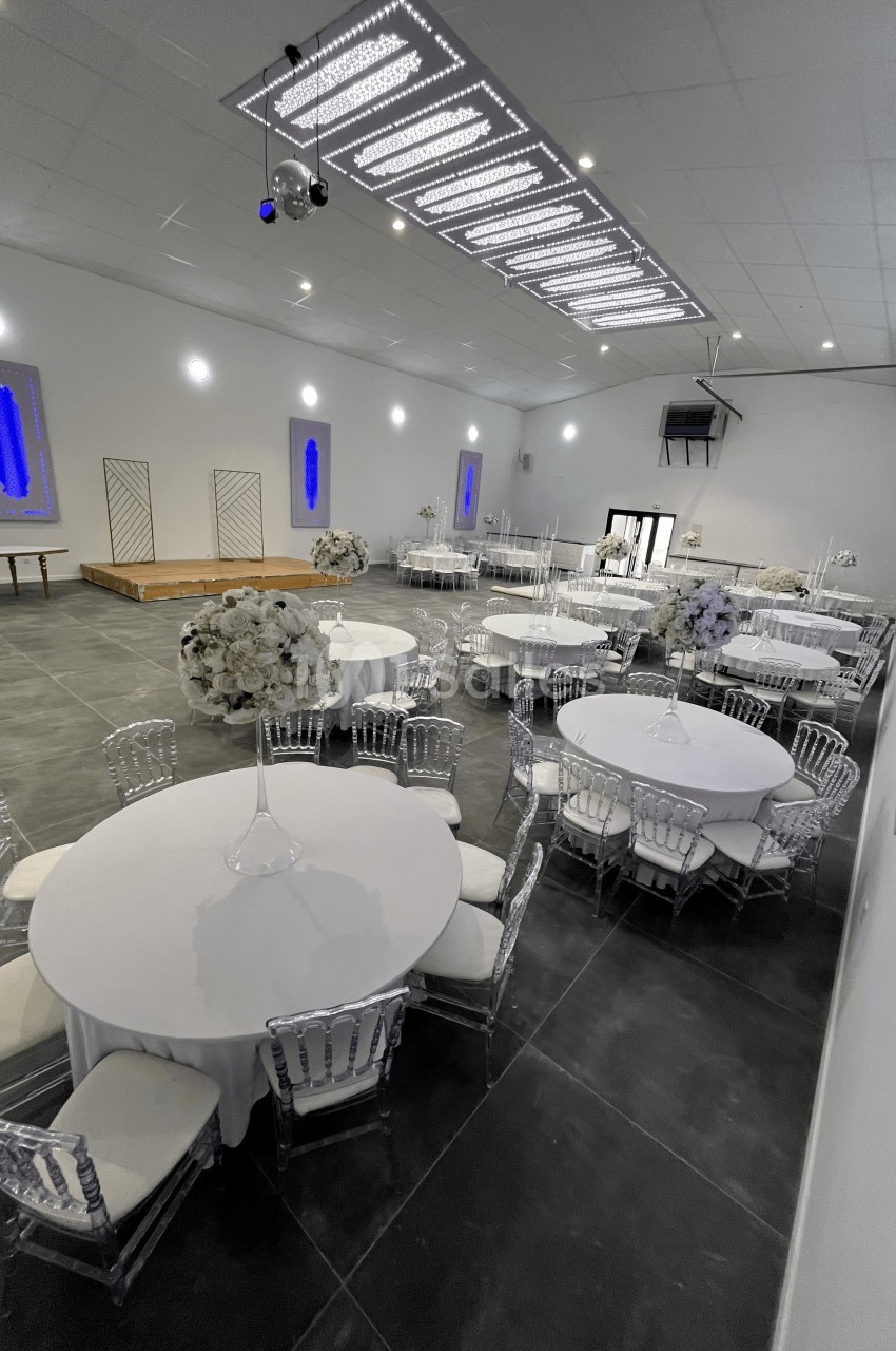 Salle de réception moderne avec tables rondes dressées, chaises transparentes et décorations florales blanches.