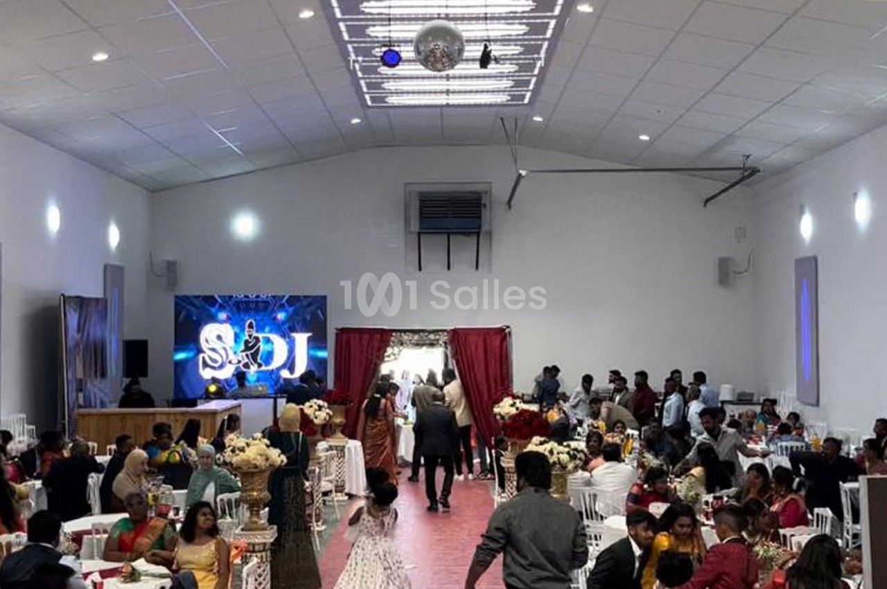 Salle de réception animée avec des invités assis et debout, décorations festives et écran lumineux en arrière-plan.