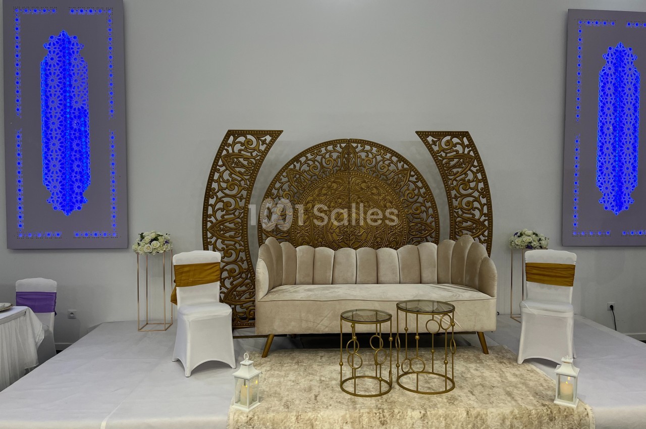 Canapé beige avec décorations dorées, tables basses et chaises blanches dans une salle ornée de panneaux lumineux bleus.