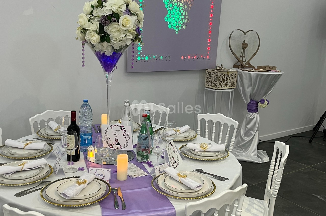 Table décorée pour un événement avec nappes blanches, chemin de table violet, vaisselle élégante et centre de table floral.