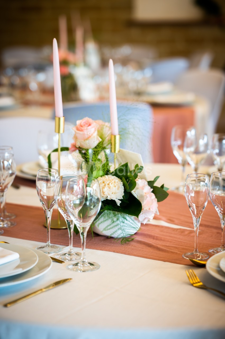 Centre de table floral avec roses et bougies roses sur une nappe blanche, entouré de verres et assiettes.