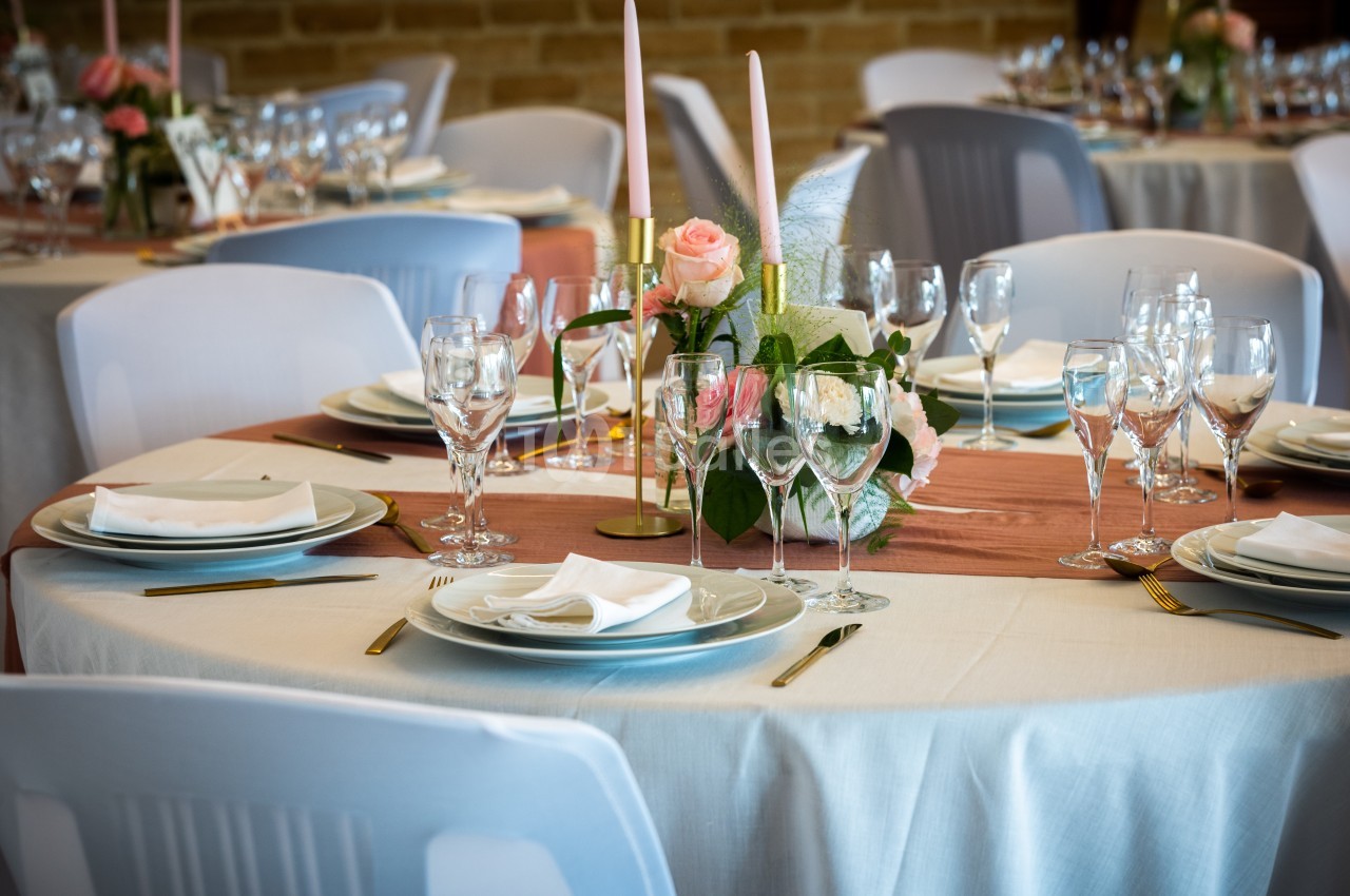Table dressée avec nappes blanches, vaisselle élégante, bougies et centre de table floral dans une salle de réception.