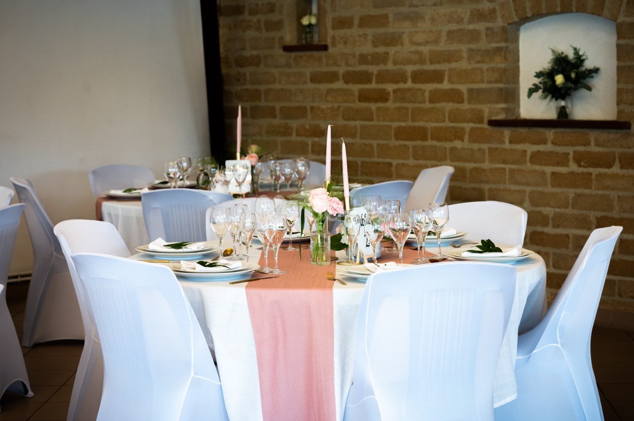 Salle décorée pour un repas, avec tables rondes dressées, nappes blanches et roses, bougies et fleurs.