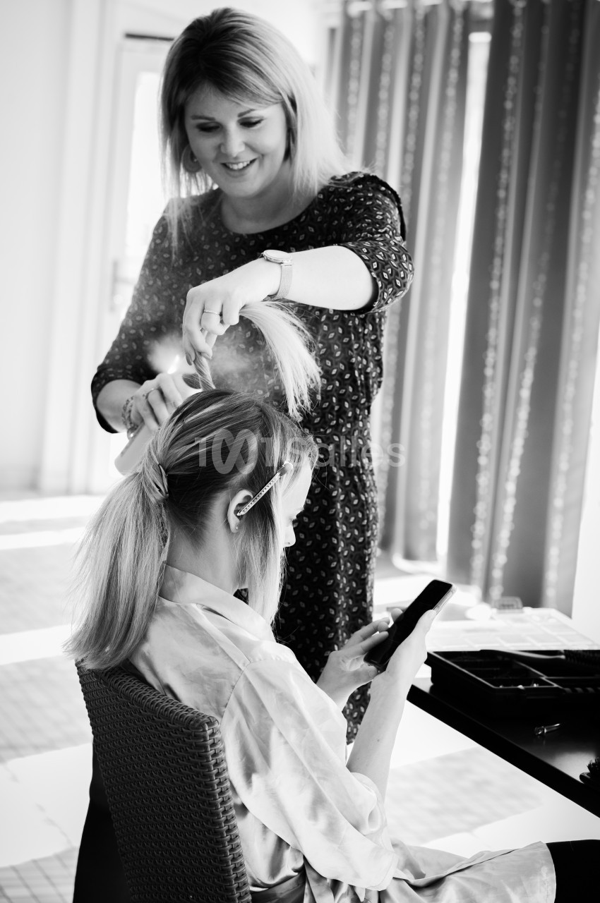 Une coiffeuse attache les cheveux d'une cliente assise, concentrée sur son téléphone, dans un salon lumineux.