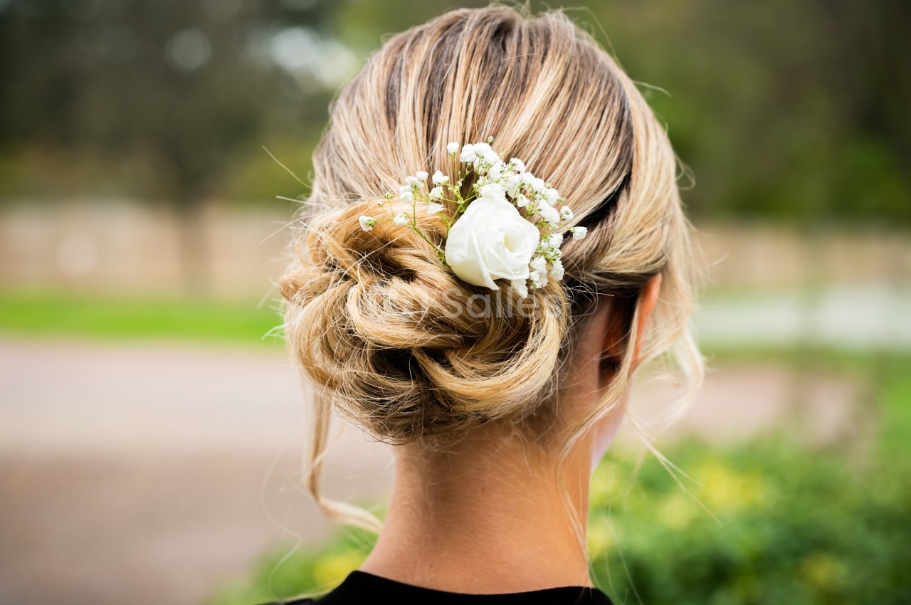 Chignon blond orné d'une rose blanche et de petites fleurs, photographié de dos dans un cadre extérieur flou.