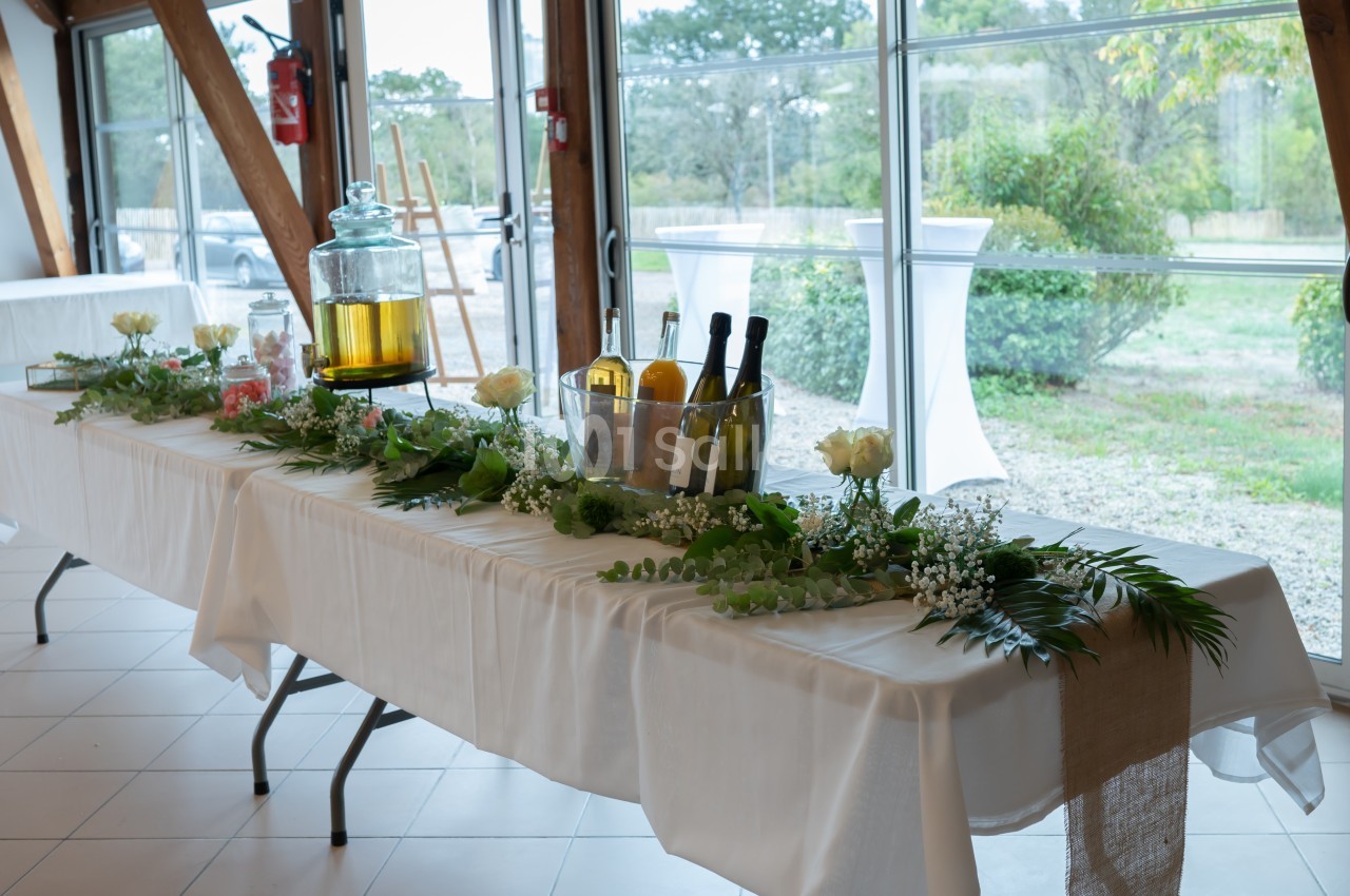 Table décorée avec nappes blanches, fleurs, feuillages, boissons et carafe, dans une salle lumineuse avec vue sur l…