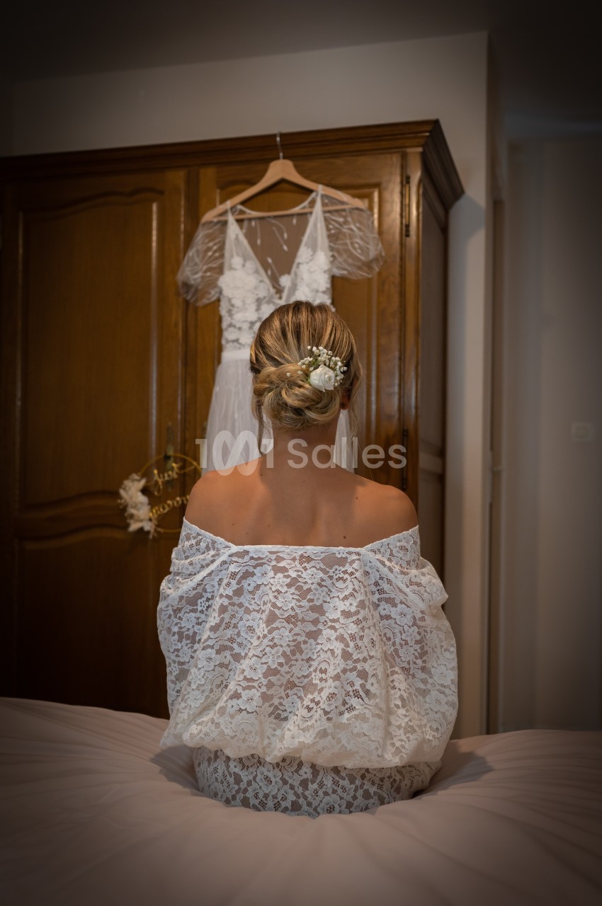 Une femme en robe en dentelle blanche, de dos, assise face à une armoire où est suspendue une robe de mariée.