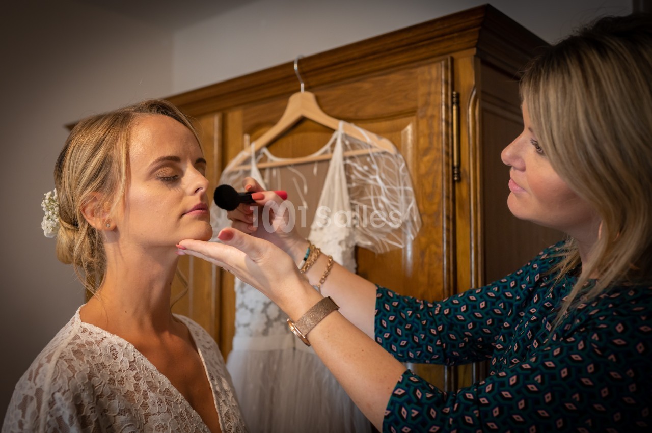 Une maquilleuse applique du maquillage sur le visage d'une femme en robe blanche devant une armoire en bois.