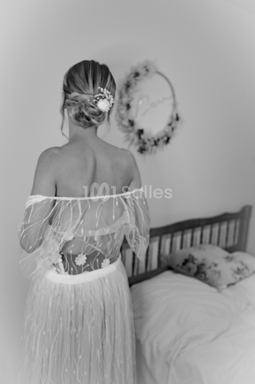 Femme de dos en robe de mariée avec un chignon décoré, debout près d'un lit dans une pièce lumineuse.