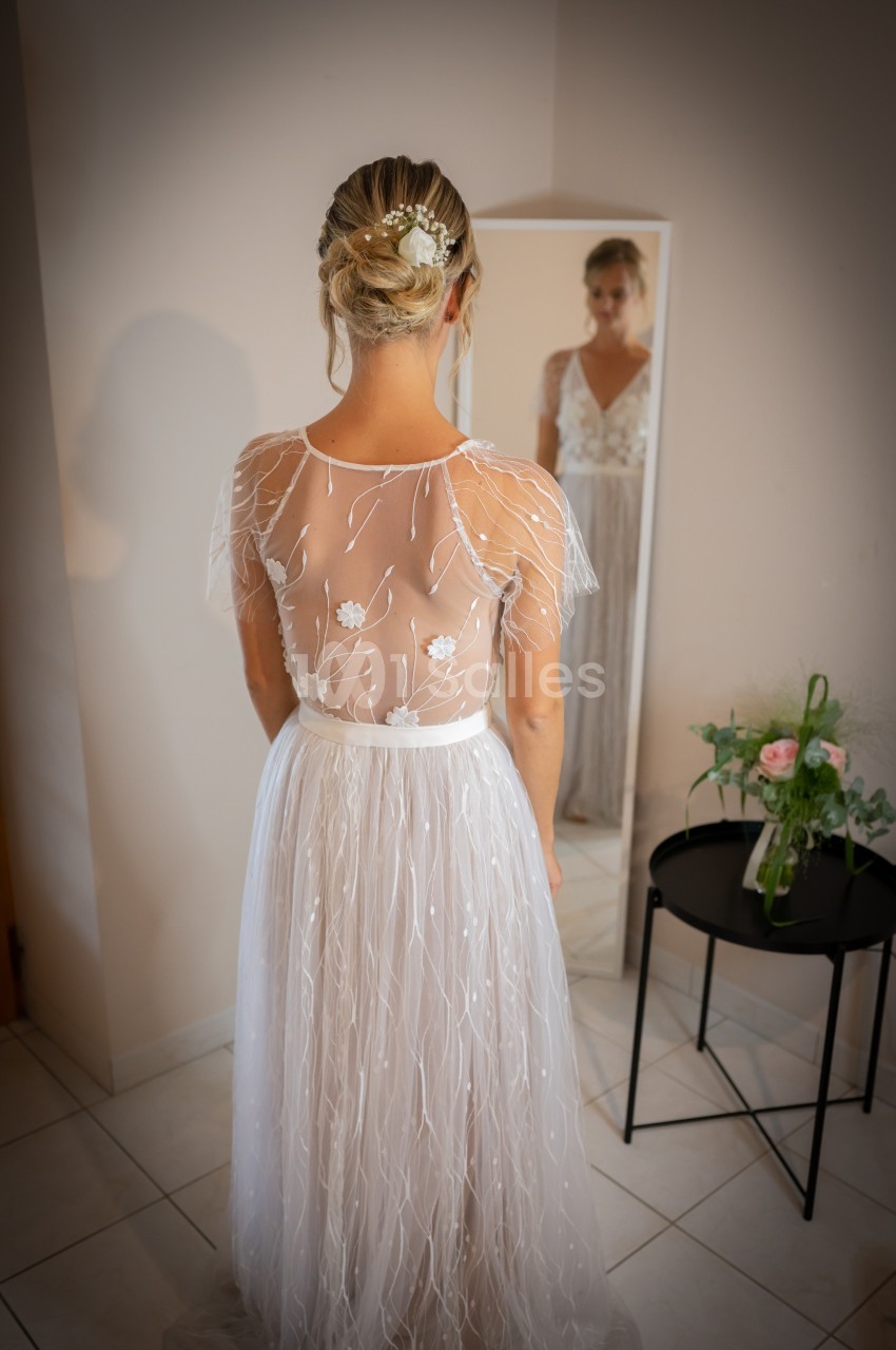 Une femme en robe de mariée vue de dos, se tenant devant un miroir dans une pièce lumineuse.