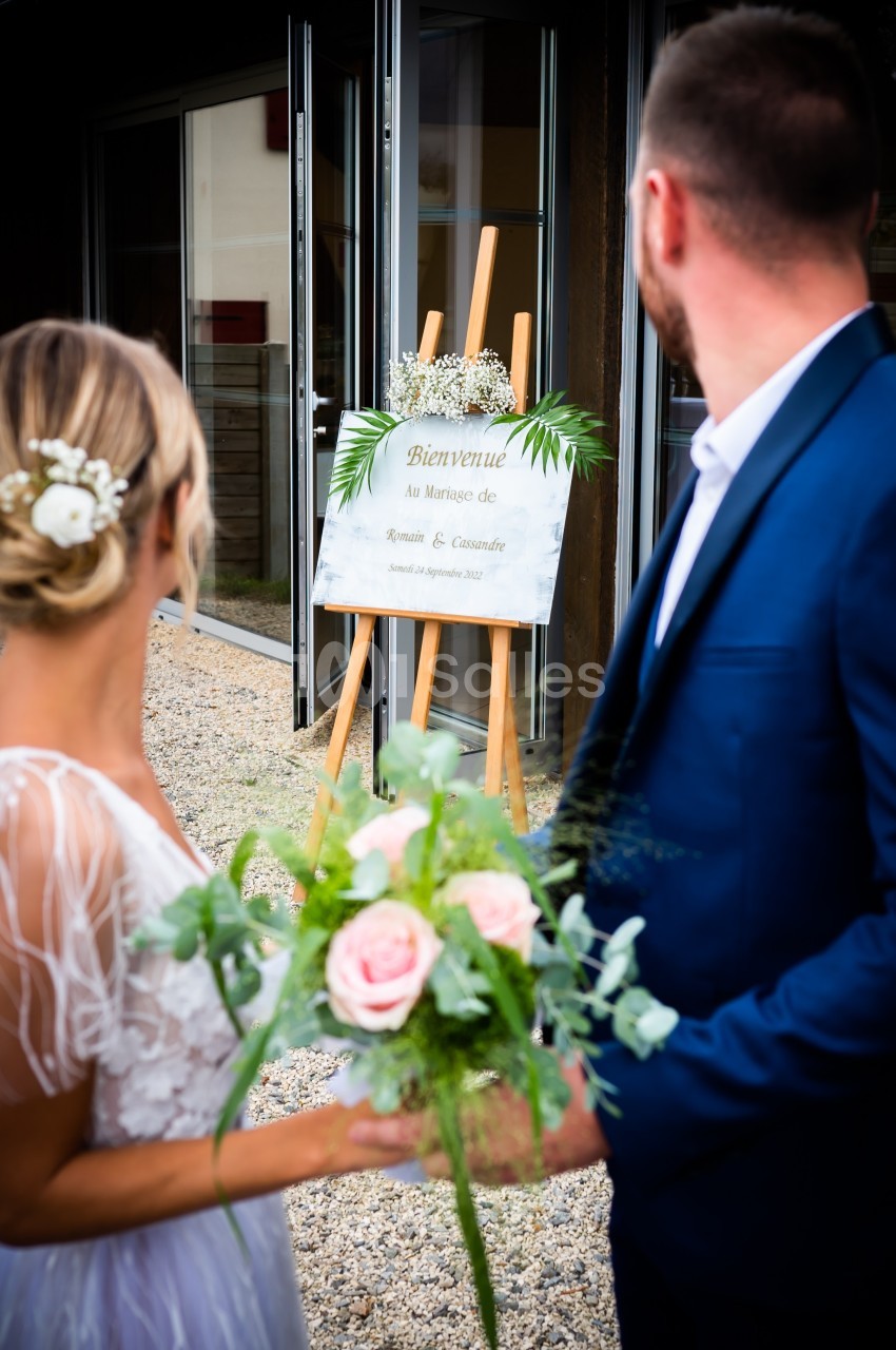 Un couple regarde un panneau de bienvenue décoré de feuillage lors d'une cérémonie de mariage en extérieur.