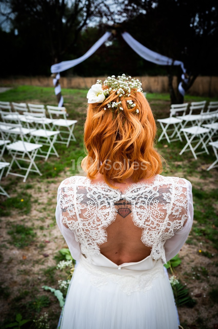 Une mariée rousse de dos, portant une robe en dentelle, face à des chaises blanches disposées en extérieur.