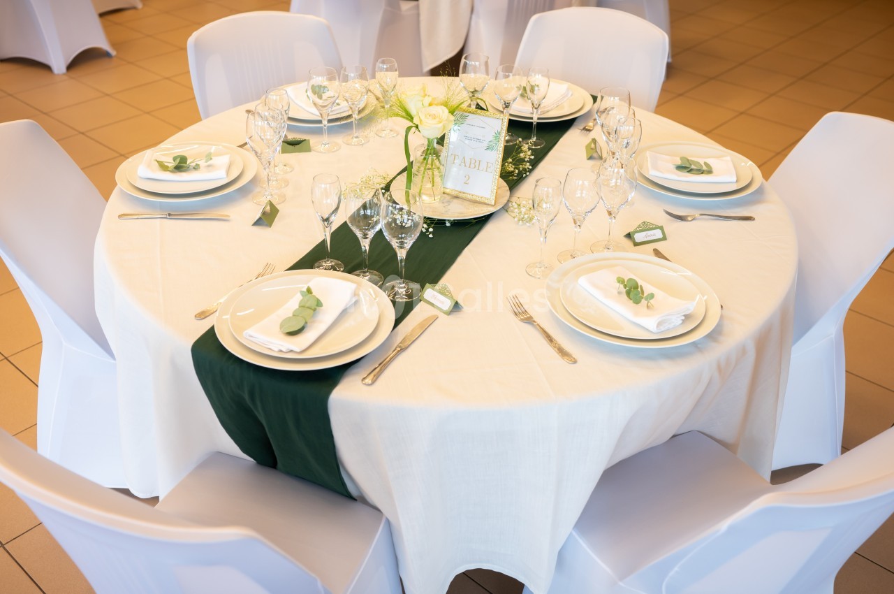 Table ronde élégamment dressée avec une nappe blanche, un chemin de table vert foncé, vaisselle et décoration florale.