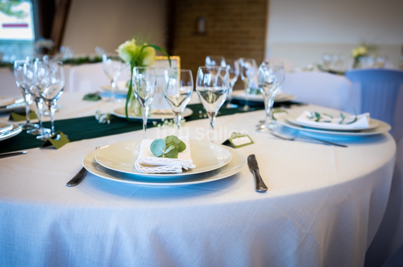 Table ronde élégamment dressée avec nappes blanches, vaisselle raffinée, verres à pied et décoration florale.