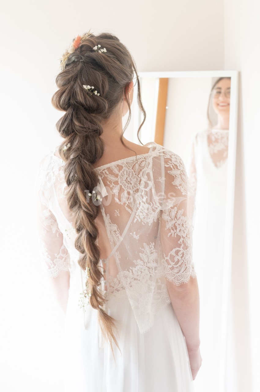 Femme en robe blanche avec une longue tresse ornée de fleurs, se regardant dans un miroir.