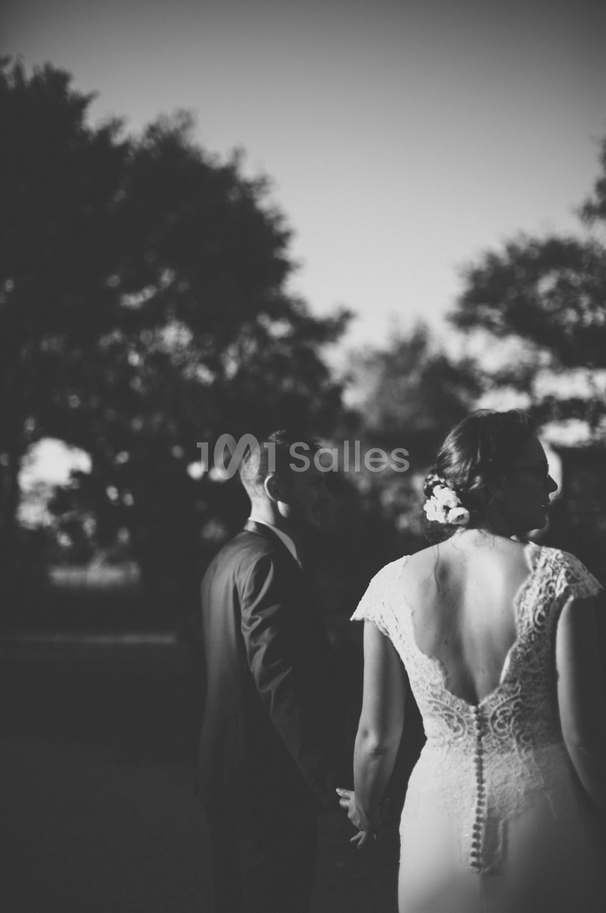 Un couple en tenue de mariage, vu de dos, se tenant la main dans un cadre extérieur en noir et blanc.