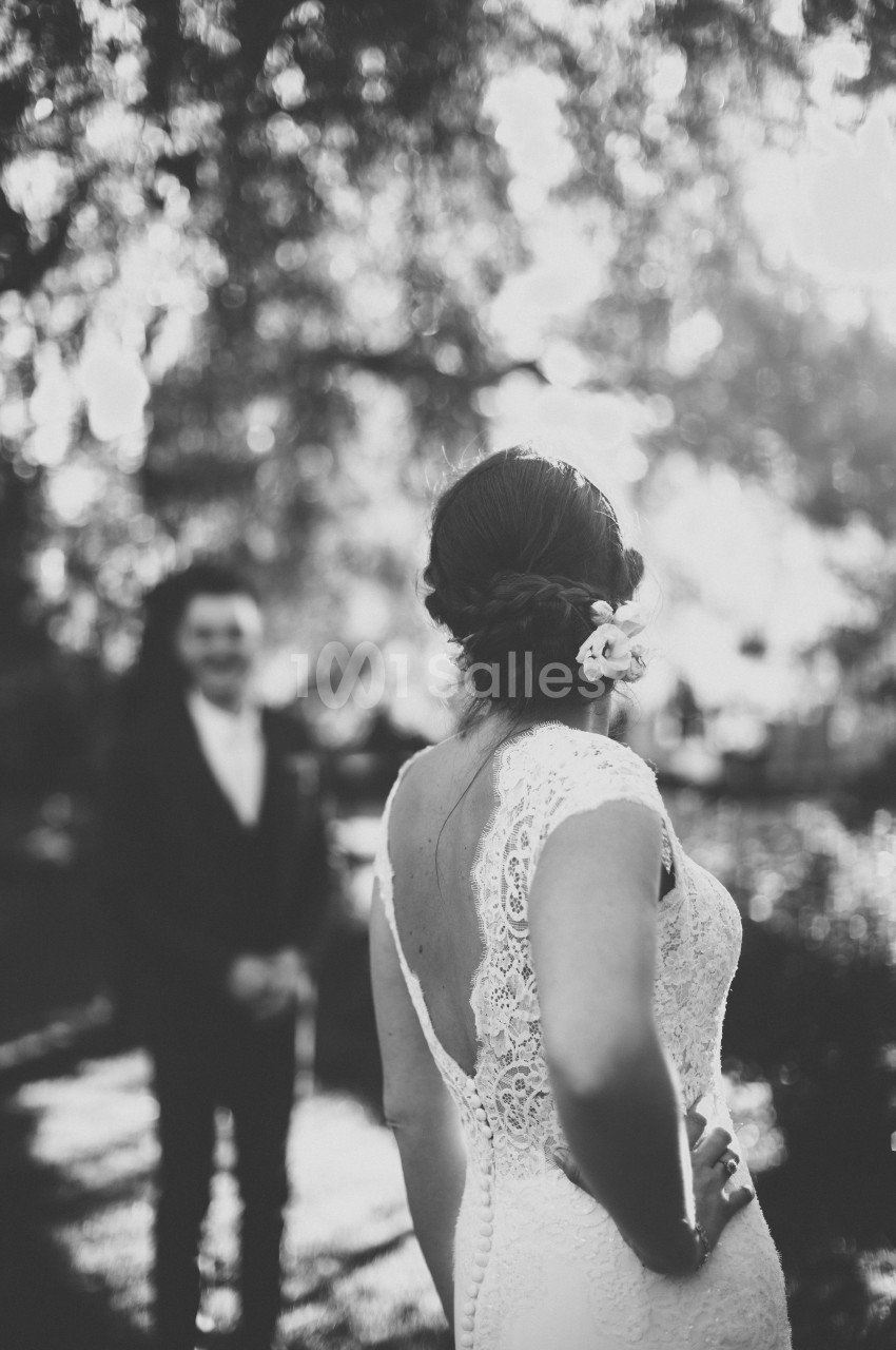 Une femme en robe de mariée vue de dos, face à un homme flou en arrière-plan dans un cadre extérieur lumineux.