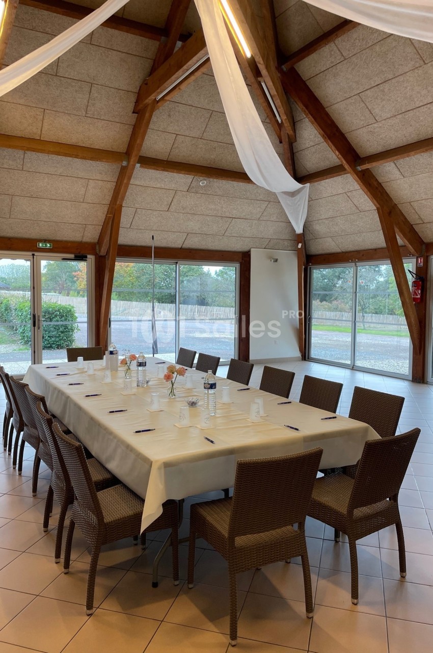 Salle lumineuse avec une grande table rectangulaire dressée, entourée de chaises en rotin, et des baies vitrées donnant sur…