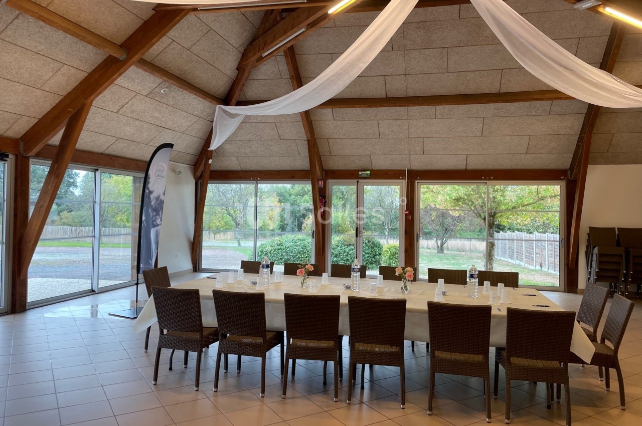 Salle lumineuse avec grande table rectangulaire dressée, chaises en rotin, baies vitrées donnant sur un jardin.