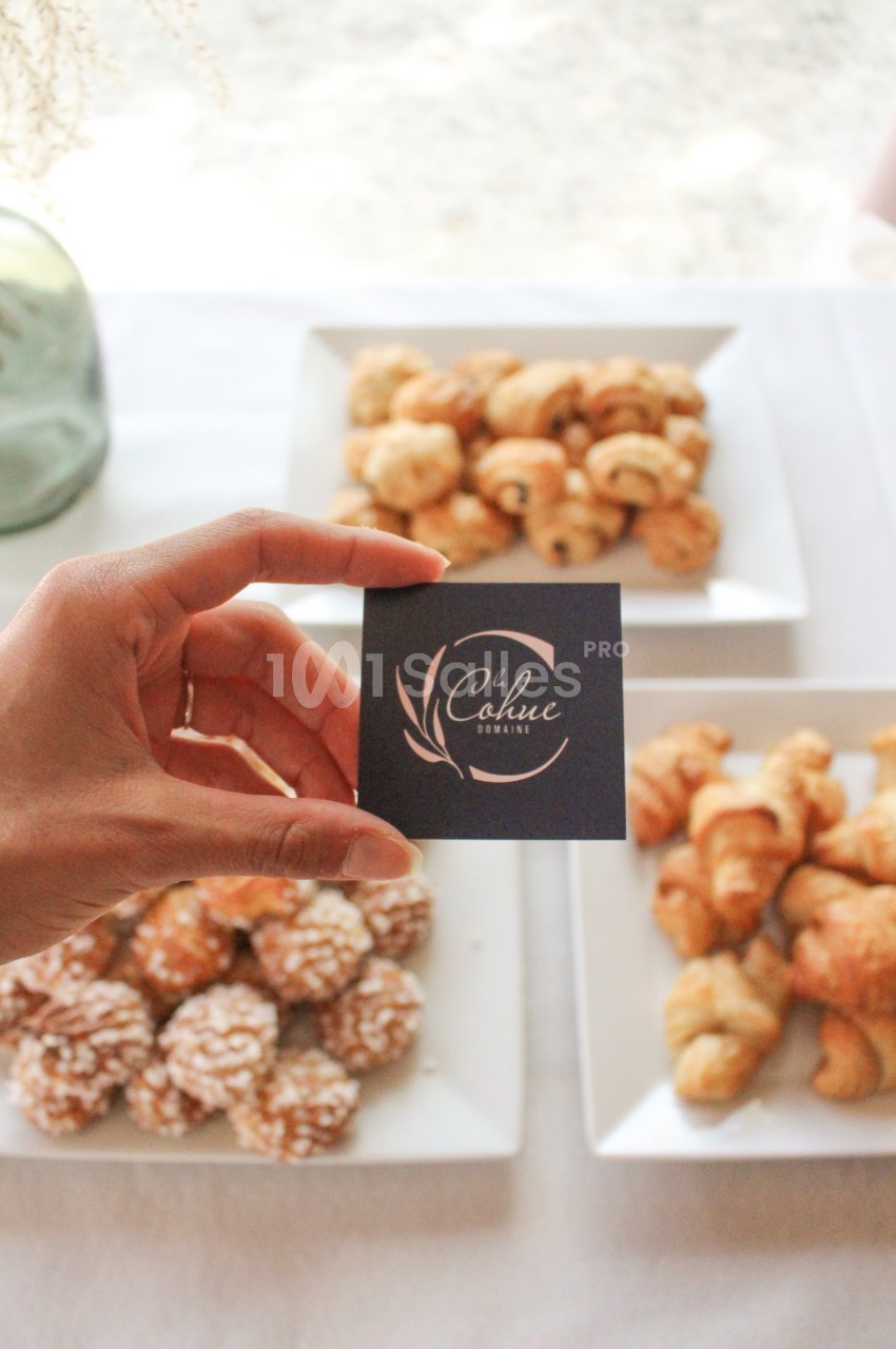 Une main tenant une carte devant des assiettes de viennoiseries variées sur une nappe blanche.