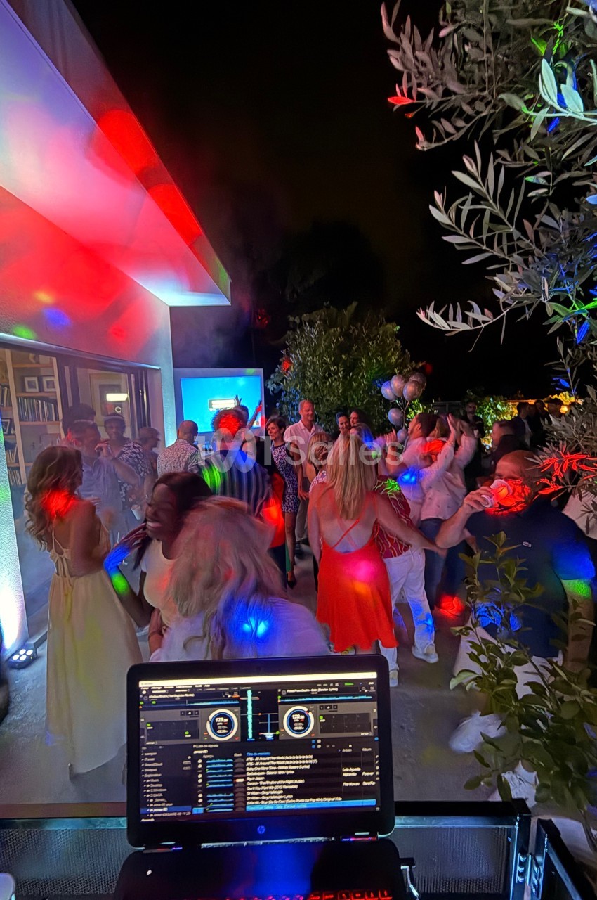 Groupe de personnes dansant en soirée en extérieur, éclairées par des lumières colorées, avec une table de mixage au premier…