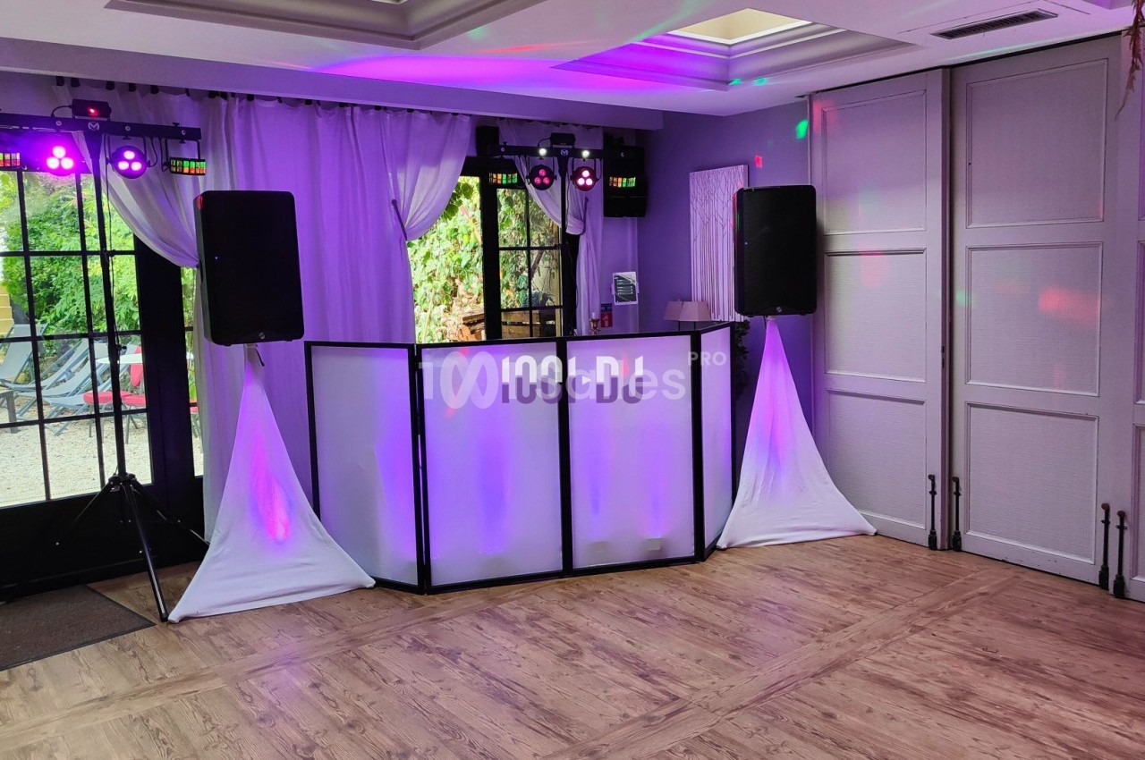 Poste de DJ installé dans une salle avec éclairage coloré, rideaux blancs et parquet au sol.