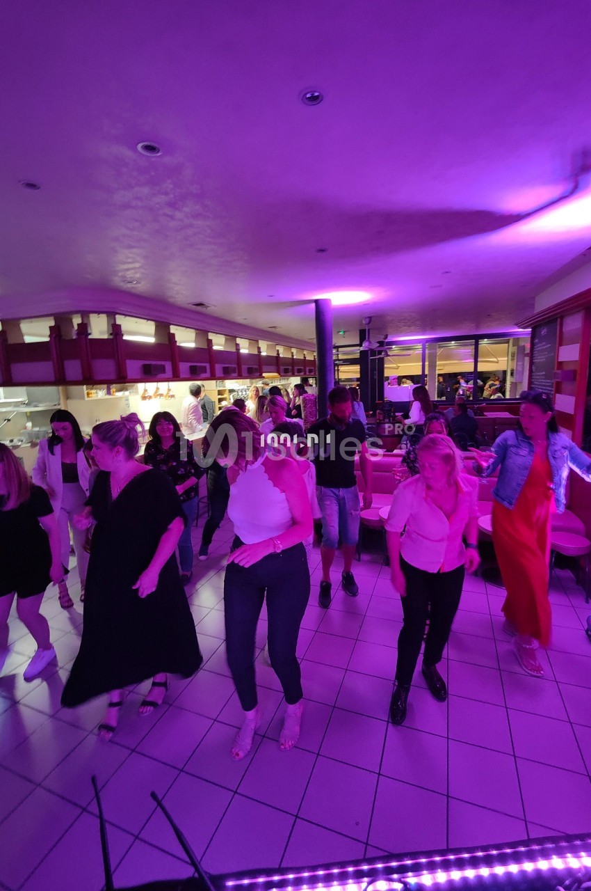 Des personnes dansent dans une salle éclairée par des lumières violettes, avec un bar en arrière-plan.