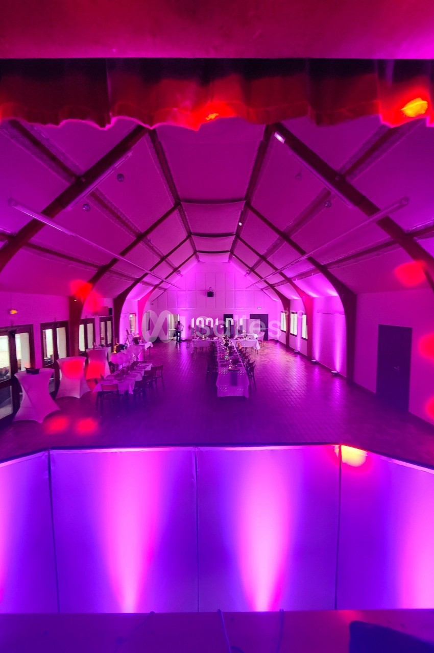 Salle de réception spacieuse éclairée par des lumières roses et violettes, avec tables dressées et décorations.
