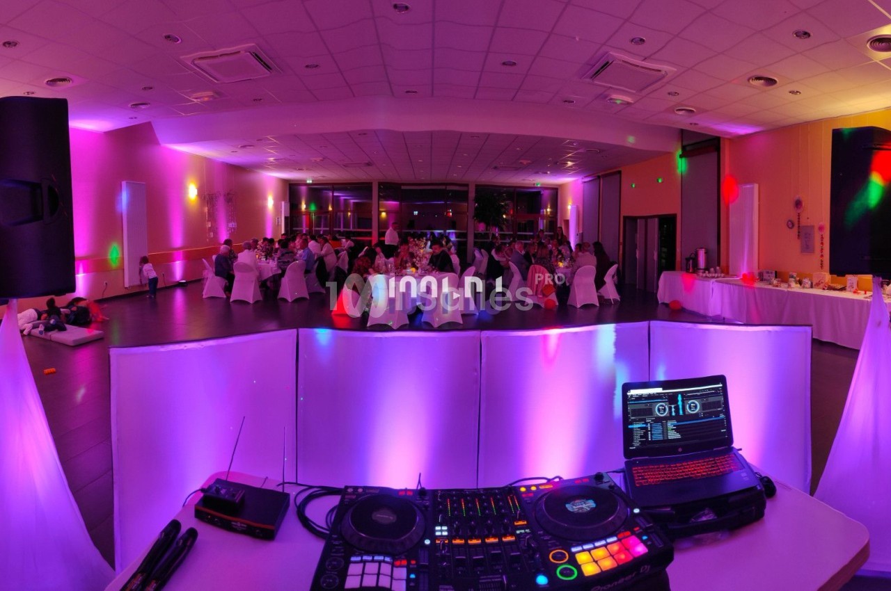 Salle de réception éclairée avec des lumières colorées, vue depuis une table de DJ équipée de matériel audio.