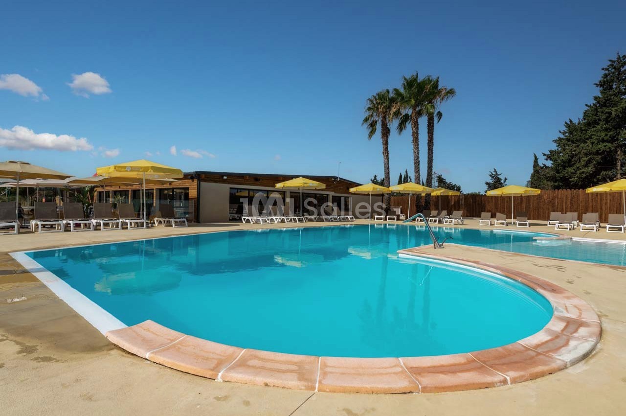 Piscine extérieure avec parasols jaunes, transats et palmiers sous un ciel bleu dégagé.