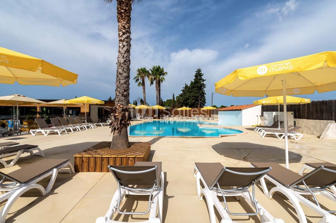 Piscine extérieure entourée de transats, parasols jaunes et palmiers sous un ciel partiellement nuageux.