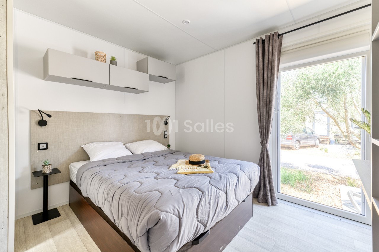Chambre lumineuse avec lit double, couvre-lit gris, étagères murales, rideaux gris et vue sur un extérieur arboré.
