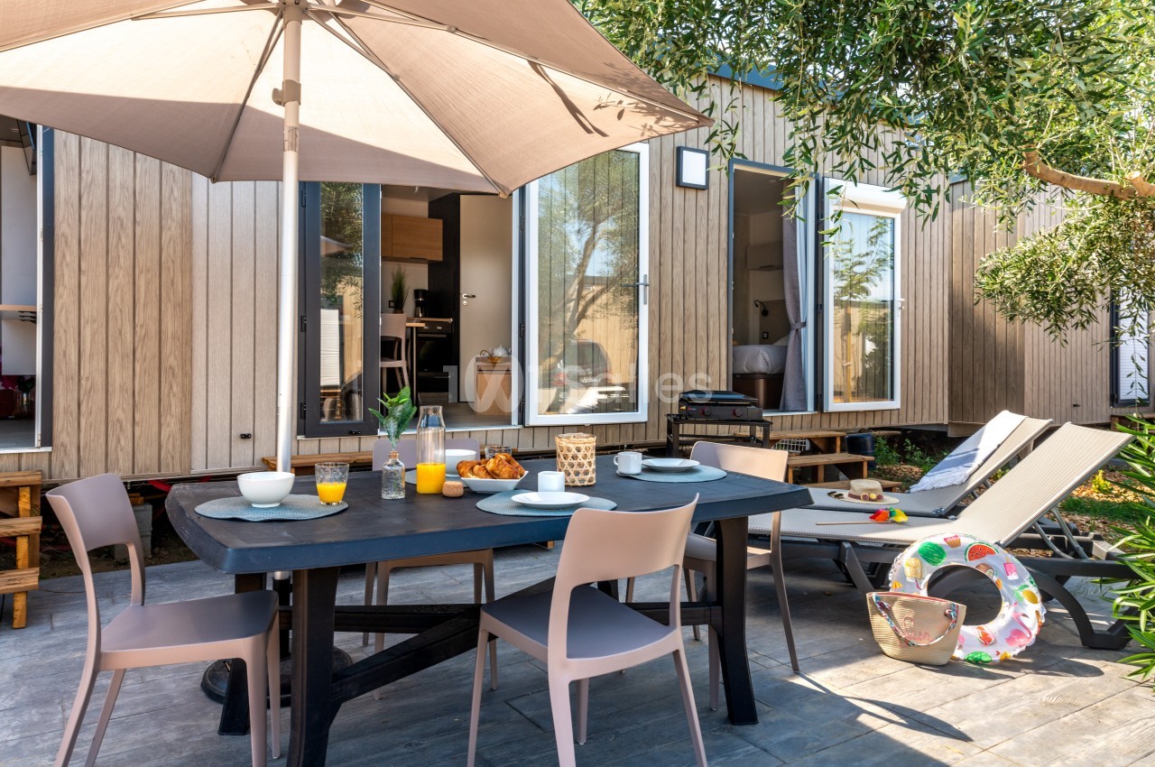 Terrasse aménagée avec table dressée pour un repas, chaises, parasol, transat et vue sur un logement moderne en bois.