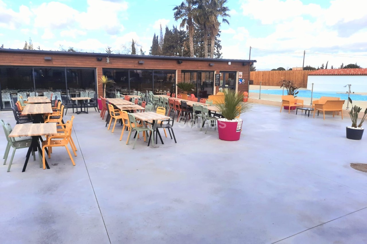 Terrasse extérieure avec tables et chaises colorées, bordée de plantes en pots, proche d'une piscine et d'un bâtiment en…