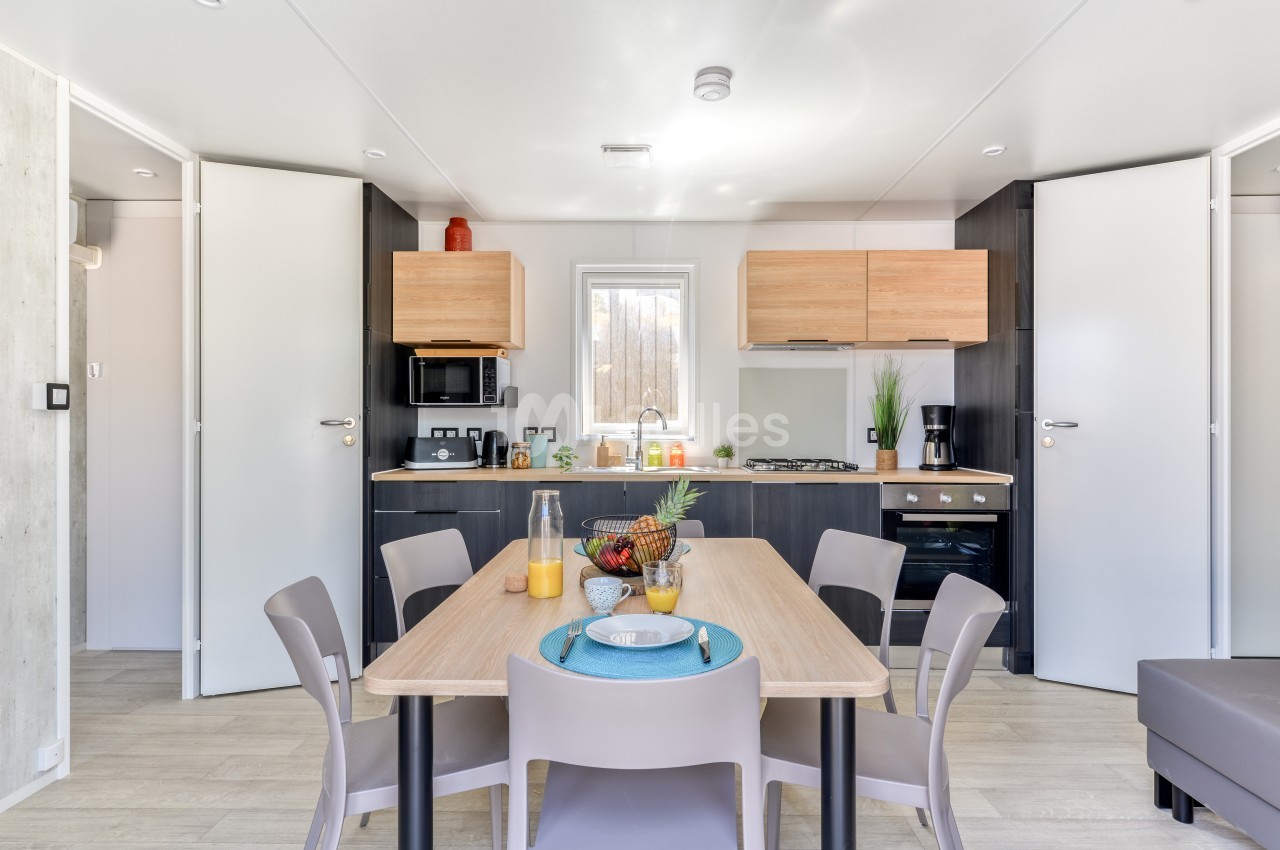 Cuisine moderne avec table à manger dressée, chaises grises, et meubles en bois clair dans un espace lumineux.