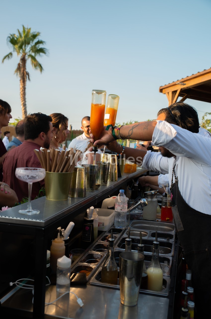 Un barman prépare des cocktails devant un groupe de personnes lors d'un événement en extérieur au coucher du soleil.