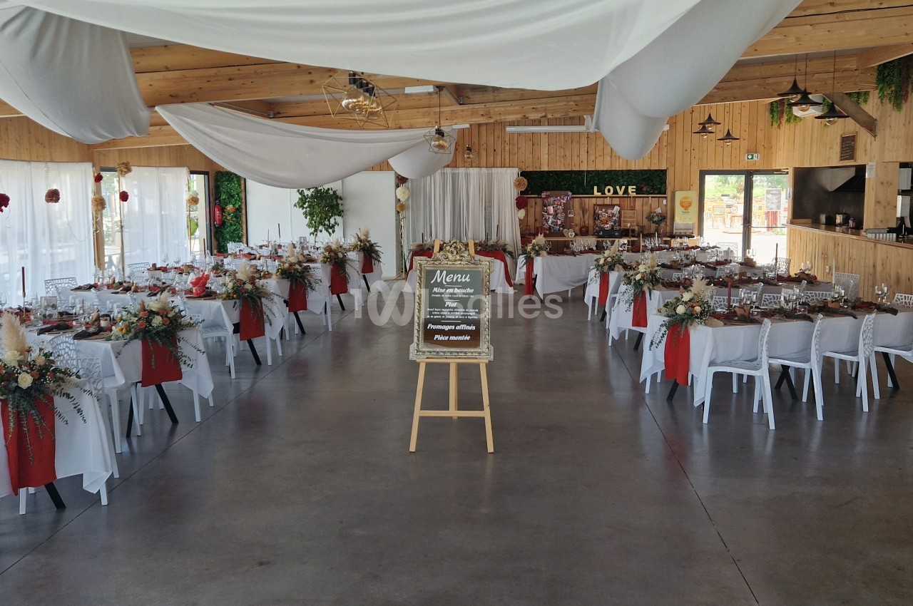 Salle de réception décorée pour un événement, avec des tables dressées, des nappes rouges et un tableau de menu au centre.