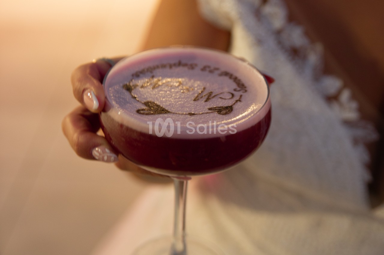 Main tenant un cocktail rose avec une mousse décorée de motifs et d'écriture, en arrière-plan flou.