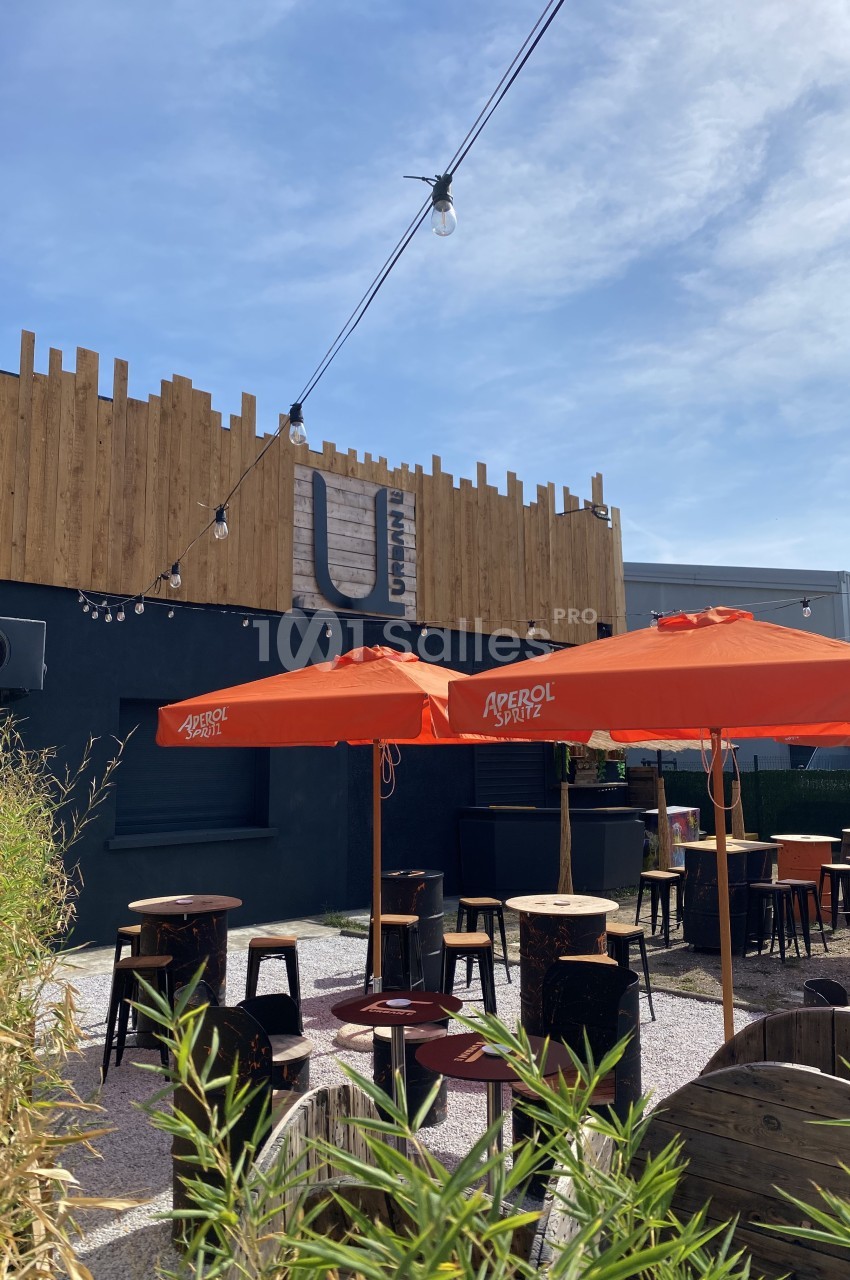 Terrasse extérieure avec tables, chaises et parasols orange, entourée de végétation, devant un bâtiment en bois et métal.