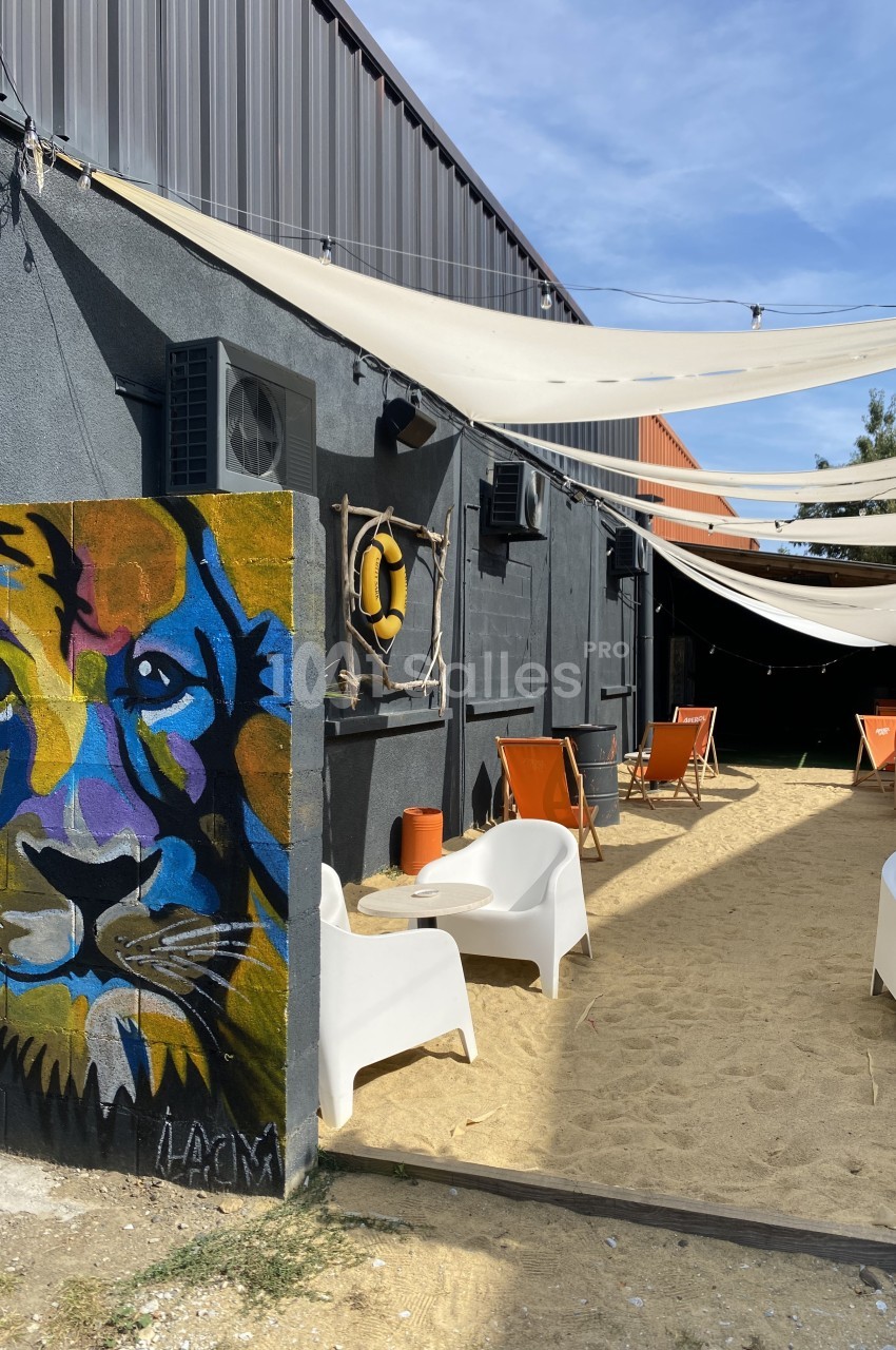 Mur peint avec un lion coloré, terrasse extérieure avec chaises longues sur du sable sous des voiles d'ombrage.