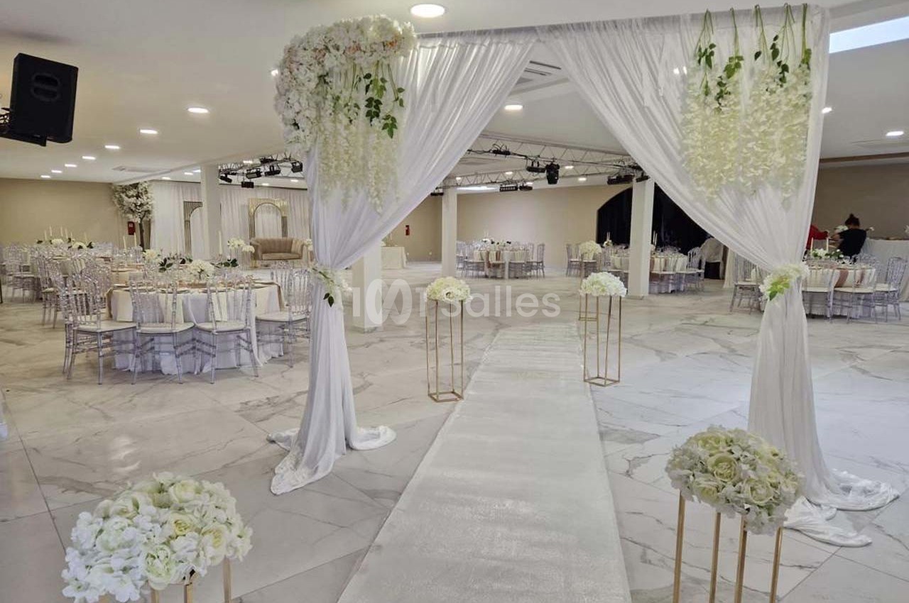 Allée centrale décorée de fleurs blanches et de rideaux, menant à une salle de réception élégamment aménagée.