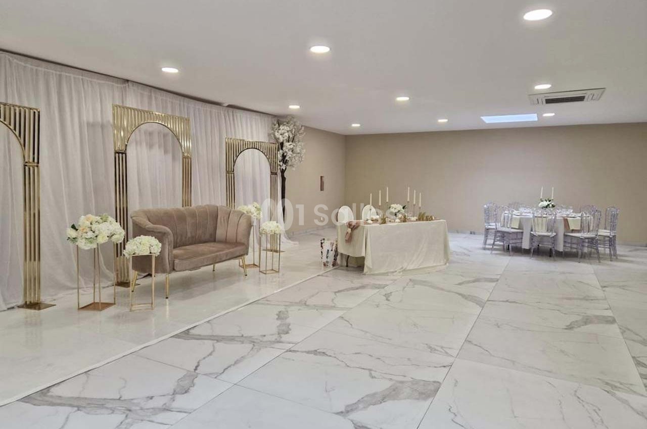 Salle de réception lumineuse avec sol en marbre, arche dorée, canapé beige et tables décorées pour un événement.