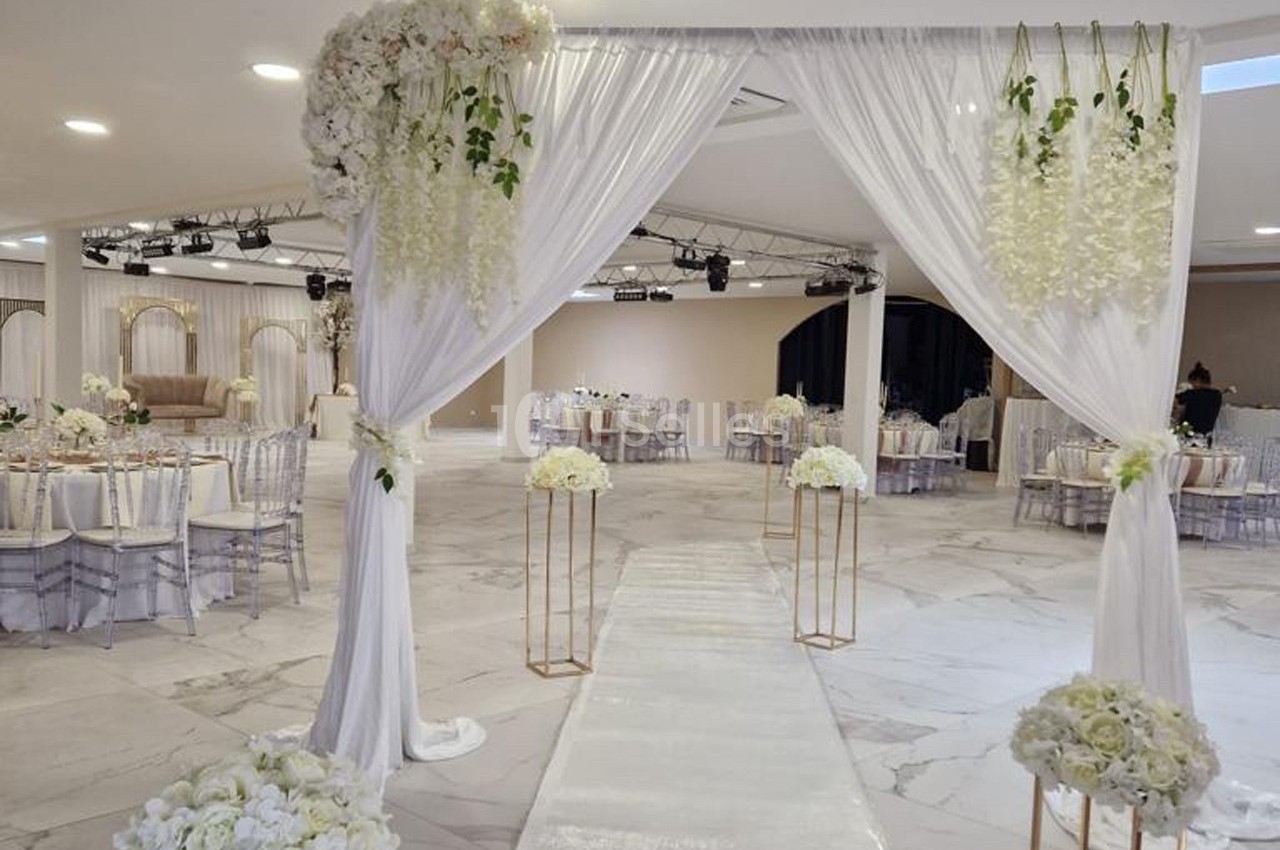 Allée décorée pour une cérémonie, avec arche florale blanche, chaises transparentes et tables dressées en arrière-plan.