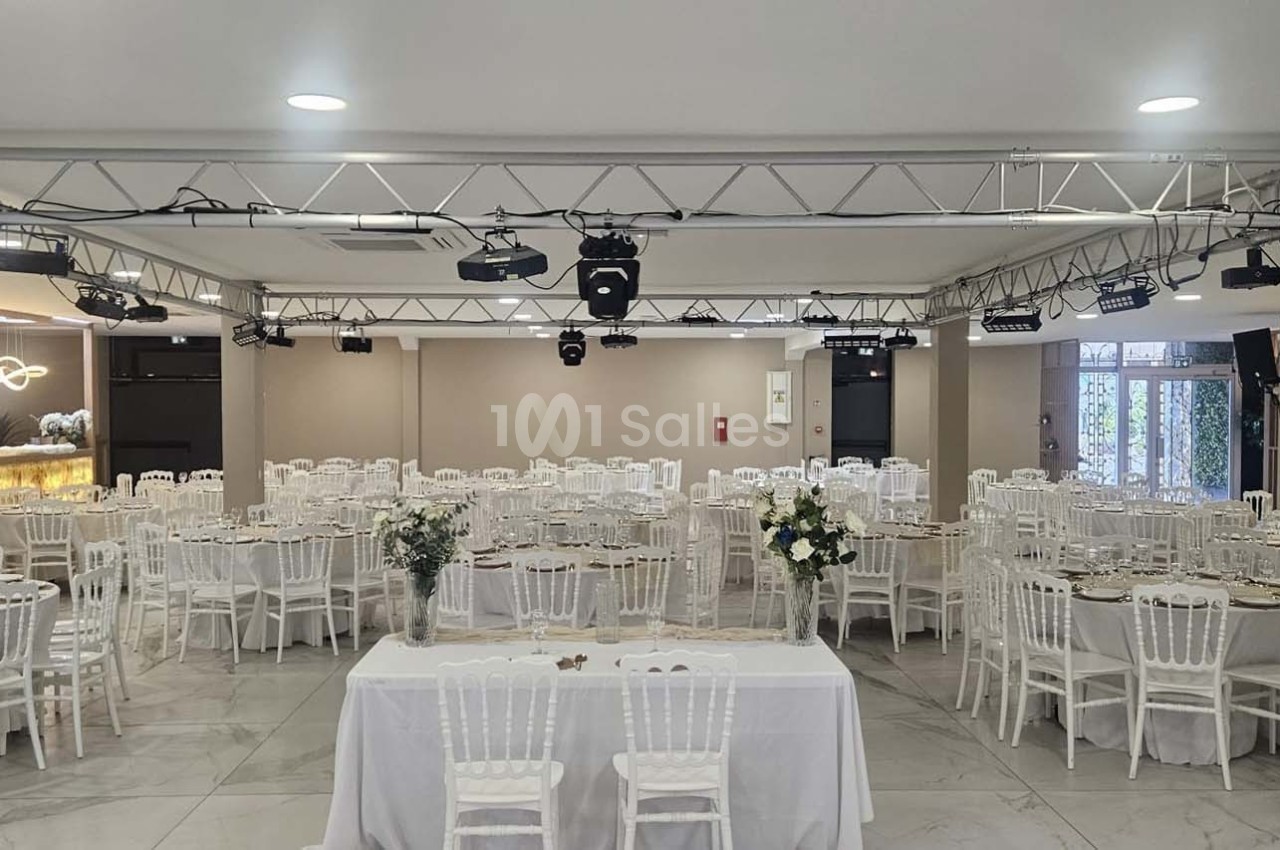 Salle de réception lumineuse avec des tables rondes dressées et des chaises blanches, décorée de fleurs.